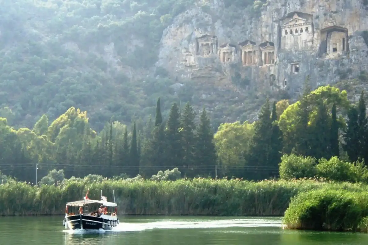 Dalyan kanalında sazlıklar arasından geçen gezi teknesi, arkada kral mezarları